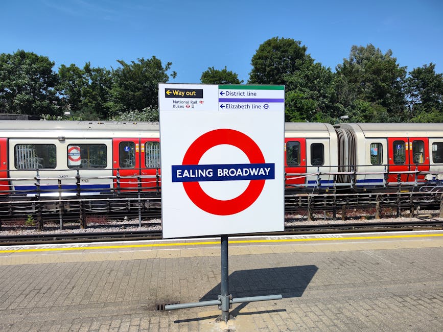 A large vertical sign at Ealing Broadway train station displaying the station name in white text on a blue background inside a red circle symbol. The sign also features directional information with arrows pointing left for 'Way out' and national rail buses, and to the right for the district and Elizabeth line services. Behind the sign, there is a stationary London Underground train with a silver body and red, white, and blue detailing on the exterior, positioned on the railway tracks. The train has rectangular windows, with some open to reveal interior seating. The platform is paved with textured concrete tiles, and a yellow safety line runs parallel to the tracks. In the background, a row of lush green trees under a clear blue sky provides a natural contrast to the industrial scene, creating a peaceful environment that subtly reflects the importance of efficient public transport and urban infrastructure. The image captures a bright day with natural lighting, emphasizing the contrast between the station's signage, the train, and the surrounding environment, illustrating a typical scene at a central railway station in London. Rubbish Clearance Ealing offers services related to waste and rubbish removal, emphasizing the importance of maintaining clean and well-organized public spaces in such busy urban areas.