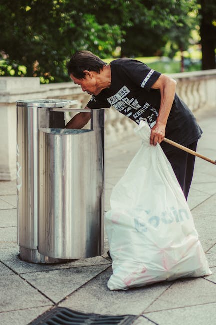 A middle-aged man with dark hair wearing a black T-shirt is seen using a wooden-handled tool to empty a large white plastic bag filled with rubbish into a cylindrical stainless steel public trash bin on a paved sidewalk. The bin has a metallic, reflective surface and appears to be designated for waste collection in an outdoor urban setting. The background includes a stone balustrade and lush green trees, suggesting a park or residential area. The man’s posture indicates he is focused on properly disposing of waste, with his right hand holding the bag open and his body slightly bent forward. Natural light illuminates the scene, highlighting the metallic finish of the bin and the textures of the man's clothing and the bag. This image illustrates an example of independent waste disposal, which is relevant to rubbish removal services like those provided by Rubbish Clearance Ealing, supporting environmentally responsible rubbish management in community spaces while showcasing practical methods for rubbish collection outside of local authority schemes.