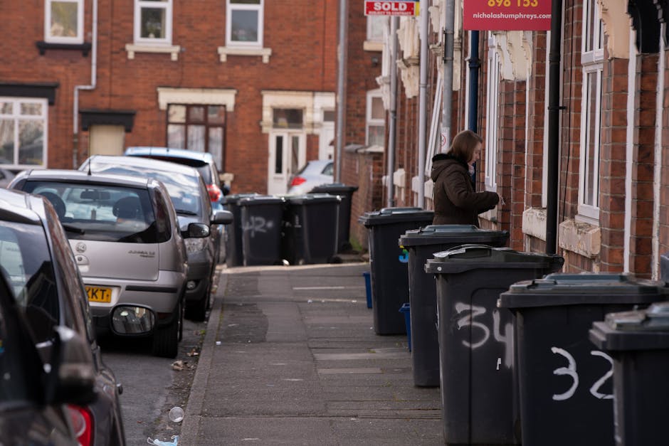 A narrow residential street with a row of brick terraced houses featuring white-framed windows and brown window sills. Several dark grey wheelie bins with white numbering are lined up along the sidewalk, some with lids partially open; a woman with shoulder-length brown hair, wearing a dark coat, is seen standing next to one of the bins, appearing to be arranging or inspecting the waste. Parked cars, including a silver hatchback at the forefront and additional vehicles further down, occupy the left side of the street, with some partially visible through the gaps between bins. The surface of the sidewalk is made of grey concrete slabs, with slight irregularities and scattered debris such as small pieces of litter. The environment appears to be during daylight with soft, natural lighting, emphasizing an ordinary scene of private waste disposal fitting within the context of alternative rubbish collection services offered by Rubbish Clearance Ealing for flats and property management. The background shows more brick houses with similar architecture and additional waste bins, indicating on-site collection or private rubbish management typical in residential areas.