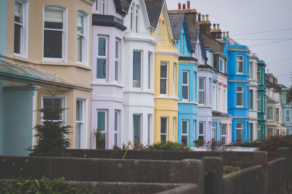 A row of terraced Victorian houses in a seaside town, each painted in pastel shades including cream, lavender, yellow, blue, and turquoise, with white window frames and decorative trim. The houses feature bay windows and pitched roofs with dark tiles, with chimneys visible on the rooftops. In the foreground, a concrete boundary wall and a small garden area with plants and shrubs separate the pavement from the houses. Overhead, power lines are visible against an overcast sky, creating a subdued atmosphere. This scene illustrates typical residential architecture in a coastal UK setting, showcasing the vibrant exterior colours that may be associated with private property maintenance and alternative disposal approaches, relevant to rubbish clearance or estate cleanouts. The neat appearance and timeless design are characteristic of properties that often require regular rubbish management or exterior cleaning services, such as those provided by Rubbish Clearance Ealing.