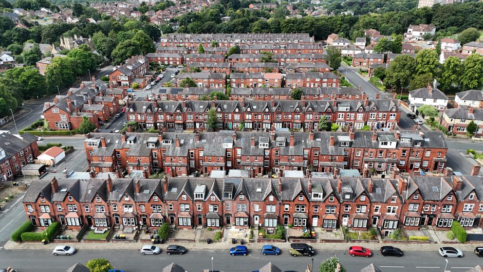 Aerial view of a residential area featuring a row of red brick terraced houses with pitched roofs and large front gardens, lined along a street with parked cars. Behind this row, there is a collection of similar red brick houses with chimneys, situated within a densely built estate. The buildings are surrounded by green spaces, trees, and other housing developments extending into the distance. The scene appears well-maintained with tidy street parking and neatly landscaped front yards. The photograph captures a clear, daytime atmosphere with natural lighting, showcasing the uniformity and density of Victorian-style terraced housing, which may be relevant to private property or estate rubbish clearance services in a UK context, such as those provided by Rubbish Clearance Ealing.