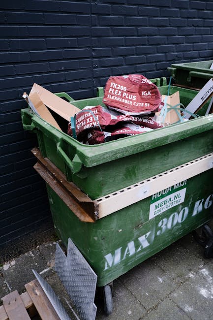 A green wheeled rubbish bin filled with red-bagged garden composting materials, such as mulch or soil amendments, is positioned against a black brick wall. The bin is placed on a paved outdoor surface, and a second similar bin is partially visible beside it. A wooden pallet supports the bin, with a metal grid or grate leaning against the pallet's side. The green bin has white printed markings indicating waste collection details, and the contents appear to be related to gardening or landscaping, typically associated with private waste management or alternative rubbish disposal methods. The scene is well-lit by natural daylight, emphasizing the textures of the plastic bin, bags, and the brick wall background, illustrating typical external refuse storage for garden waste or miscellaneous household rubbish suitable for independent collection services, such as those offered by Rubbish Clearance Ealing.