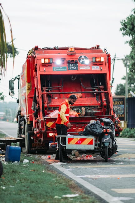A commercial waste collection worker in a high-visibility yellow and orange jacket is operating a large red refuse collection vehicle parked on the side of a paved urban street. The back of the vehicle is open, revealing mechanical components and hydraulic arms used for bin lifting. The worker is leaning slightly forward, loading a black garbage bag into the rear compartment. Nearby, a small black motorbike is parked, partially obstructed by a metal barrier with reflective red and white stripes, which is positioned on the curb to prevent vehicle encroachment. The scene is set in a residential or commercial area with a green grassy verge and some trees in the background, along with a few electrical wires overhead. The lighting suggests daytime, with a slightly overcast sky diffusing natural light evenly across the environment. The image captures the practical scene of on-site waste removal, highlighting the efforts of private rubbish collection services like Rubbish Clearance Ealing in managing waste outside of standard municipal collection schedules, emphasizing the operational details involved in efficient rubbish removal and disposal.
